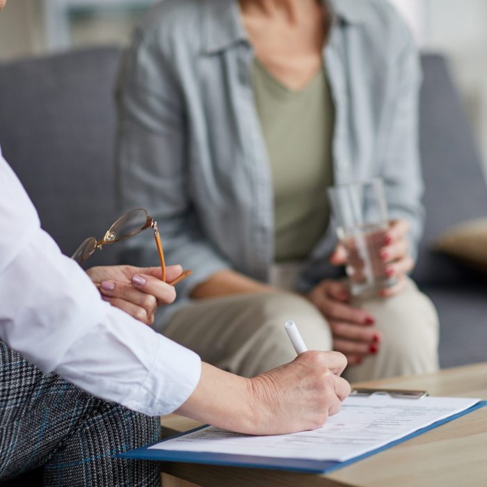 Close up of unrecognizable female psychologist writing on clipboard while talking to mature woman during therapy session, copy space